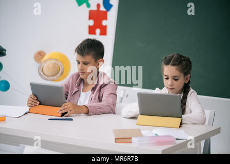 Concentrated schoolboy and schoolgirl using digital tablets while sitting at desk in classroom Stock Photo