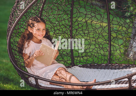 smiling african american girl reading book while sitting in swinging hanging chair in park Stock Photo