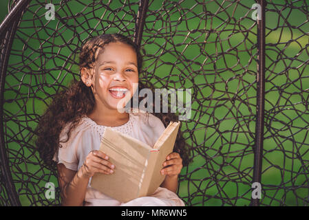 smiling african american girl reading book while sitting in swinging hanging chair Stock Photo