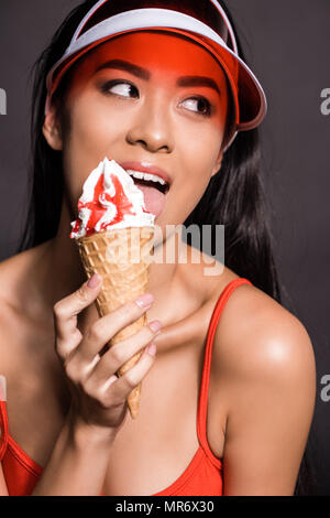 Portrait shot of beautiful asian woman in orange swimsuit and visor, licking ice-cream and looking aside Stock Photo