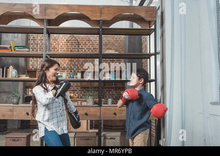 Young woman with punching pads on hands and her little son in boxing gloves having fun and laughing in living room at home Stock Photo