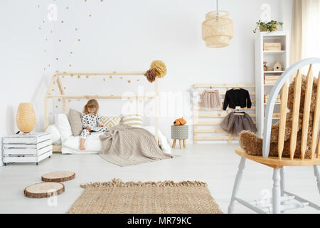 Little girl sitting on her bed in white scandinavian bedroom Stock Photo