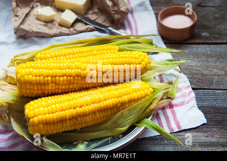 Close up tasty boiled sweet corn cobs with leafs on plate, butter and pink salt on the white napkin on the wooden rustic tabe. Selective focus Stock Photo