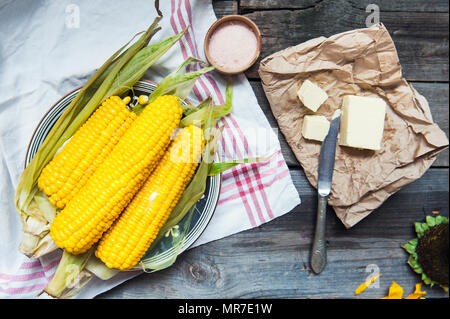 Close up tasty boiled sweet corn cobs with leafs on plate, butter and pink salt on the white napkin on the wooden rustic tabe. Selective focus Stock Photo