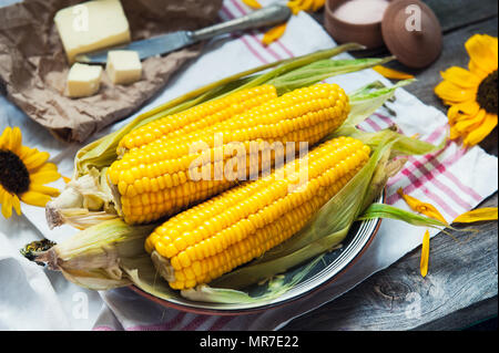 Close up tasty boiled sweet corn cobs with leafs on plate, butter and pink salt on the white napkin on the wooden rustic tabe. Selective focus. Stock Photo