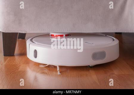 Robotic vacuum cleaner runs under sofa in room on laminate floor. Robot controlled by voice commands to direct cleaning. Modern smart cleaning technol Stock Photo