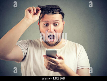 Super amazed young man with overweight reading shocking news on smartphone holding glasses up with mouth opened. Stock Photo