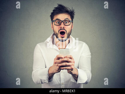 Young handsome bearded man in shirt holding new smartphone and looking extra shocked at camera. Stock Photo