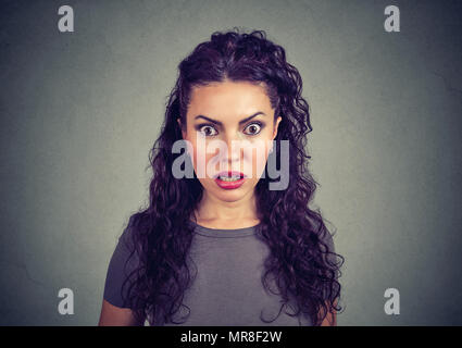 Young stunned brunette with curls holding eyes wide opened and looking scared at camera on gray. Stock Photo