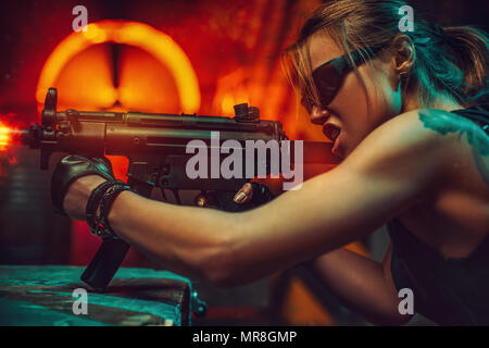 Young strong woman warrior shooting from machine gun in dramatic urban night scene. Tattoo on body. Stock Photo