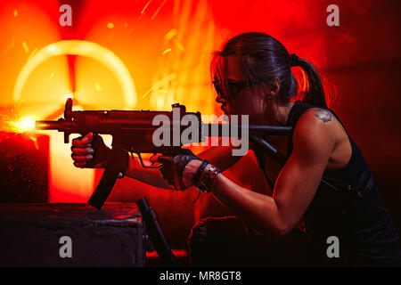 Young strong woman warrior shooting from machine gun in dramatic urban night scene. Tattoo on body. Stock Photo