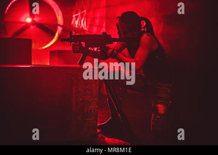 Young strong woman warrior shooting from machine gun in dramatic urban night red colors scene. Tattoo on body. Stock Photo