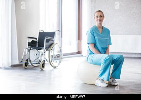 Young female physiotherapist sitting on Swiss ball with wheelchair in background. Stock Photo