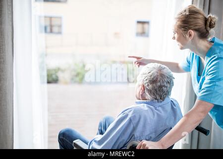 Senior man in wheelchair with care worker looking out of window. Stock Photo