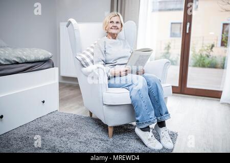 Senior woman sitting in armchair with magazine in care home. Stock Photo