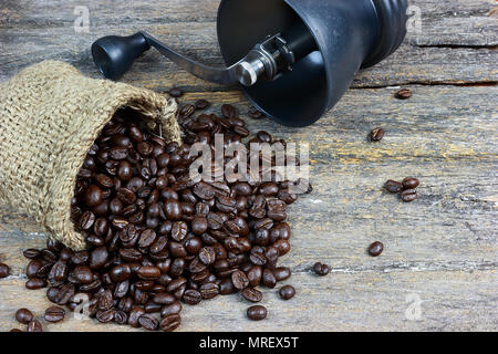 Roasted coffee beans , packed in small hemp sack, poured on a wooden board. Have a coffee grinder with hands on the background. Stock Photo