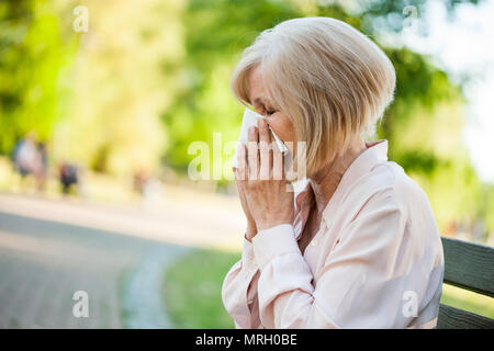 Adult woman is sitting in park and blowing nose. She is having allergy. Stock Photo