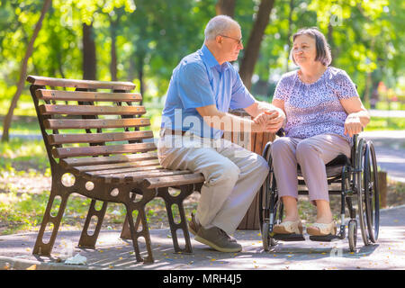 Elderly man strengthens his wife in the wheelchair outside the summer park. Stock Photo