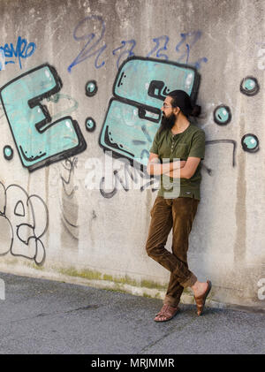 Full length portrait of young adult bearded Indian man posing against graffiti wall, looking away from camera. Stock Photo