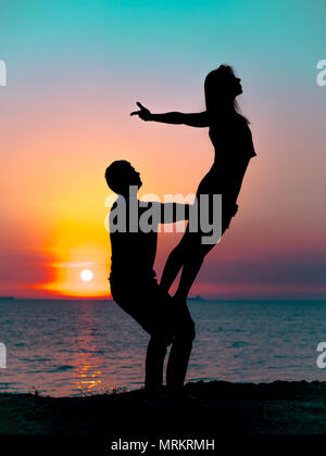 Silhouette of young woman and man doing pair yoga on sea beach at sunset. Meditation. Couple practicing acro yoga. Flexibility workout at nature background. Stock Photo
