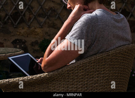 Sunlit side view profile of suntanned mature woman with reading glasses sitting in wicker garden chair on patio looking at iPad screen Stock Photo