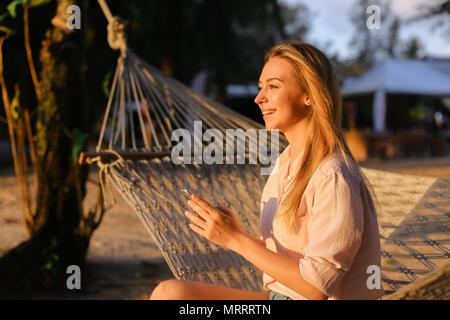 Young smiling woman using smartphone and sitting in wicker hammock on tropical resort. Stock Photo