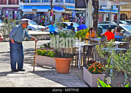 An old local Greek man with walking stick looks at people relaxing in a harbourside café in the seaside port of Sami, Kephalonia / Cephalonia Stock Photo