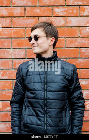 a young man stands leaning against a brick wall and smiling. Stock Photo