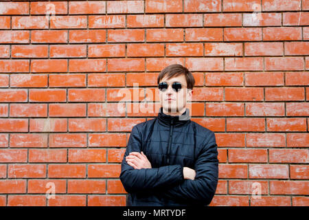 a young man stands leaning against a brick wall in sunglasses cross his arms. Stock Photo