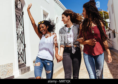 Three women wearing fashionable clothes walking on street on a sunny day. Young woman jumping in joy while walking with friends on a street. Stock Photo
