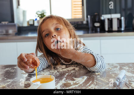Innocent young girl breaking an egg into a small pan  and tasting the batter. Cute young girl baking in the kitchen. Stock Photo
