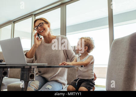 Little boy sitting with her mother talking on cell phone. Busy woman with son in home office. Stock Photo