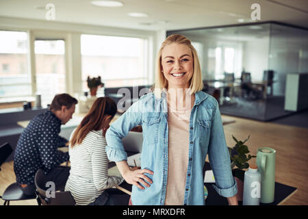 Young businesswoman smiling confidently while standing in an office with coworkers sitting in the background Stock Photo
