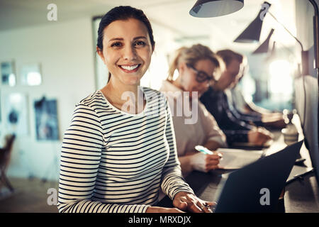 Young designer smiling confidently while working on a laptop at a table with colleagues in a modern office