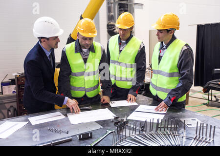 Engineer, foreman and workers wearing safety helmets are meeting at factory and discussing papers and bluprints at the table Stock Photo