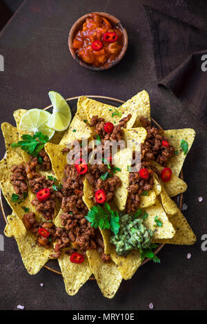 Mexican nacho corn tortilla chips with cheese, meat, guacamole and red hot spicy salsa. Nachos with ground beef on dark rustic background, copy space. Stock Photo