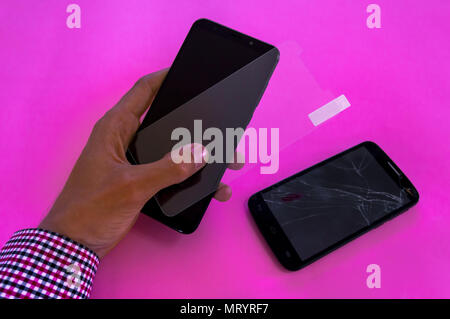 Hands of a young man holding a screen protector and a mobile phone. Broken device on pink background. Protecting a smartphone with a tempered glass. Stock Photo