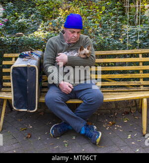 Homeless caucasian man with his small dog and his belongings sit on a yellow bench in a park image with copy space in landscape format Stock Photo