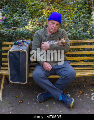 Homeless caucasian man with his small dog and his belongings sit on a yellow bench in a park image with copy space in landscape format Stock Photo