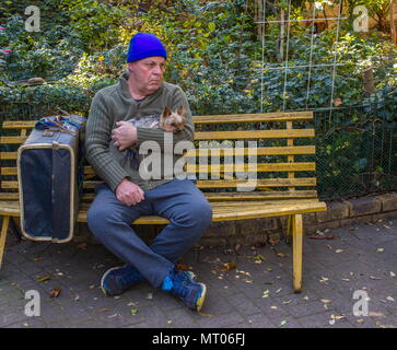 Homeless caucasian man with his small dog and his belongings sit on a yellow bench in a park image with copy space in landscape format Stock Photo