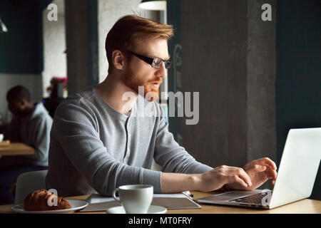 Focused millennial redhead man using laptop sitting at cafe tabl Stock Photo