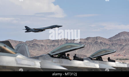 A group of F-22 Raptors from the 95th Fighter Squadron at Tyndall Air Force Base, Fla., sit on the flightline as a B-1B Lancer from the 34th Bomb Squadron at Ellsworth Air Force Base, S.D., takes off during Red Flag 17-3 at Nellis Air Force Base, Nev., July 18, 2017. Red Flag is designed to test the capability and interoperability between multiple air frames, and give the supporting personnel the chance to work together. (U.S. Air Force photo by Senior Airman Dustin Mullen/Released) Stock Photo