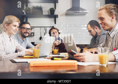 Group of cheerful coworkers sitting at table with laptops and paper documents and make brainstorming. Adult hipsters working together and discuss new  Stock Photo