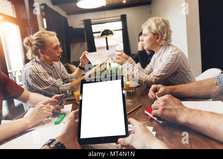 POV view of white blank screen tablet in male hands and group of young coworking people discussing new project start up in open space. Teamwork proces Stock Photo