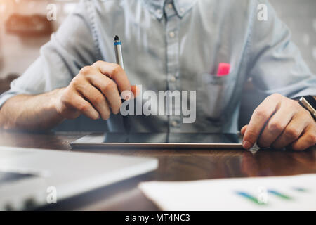 Young businessman holds a stylus in his hand and works on tablet. Tablet on a wooden table. Creative manager uses a touch pad Stock Photo