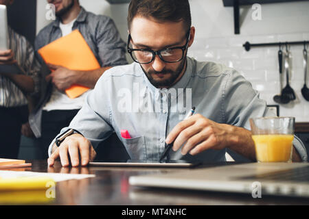 Young bearded business analyst conducts research on a computer tablet. Group of young coworkers discussing ideas with each other in modern office. Bus Stock Photo