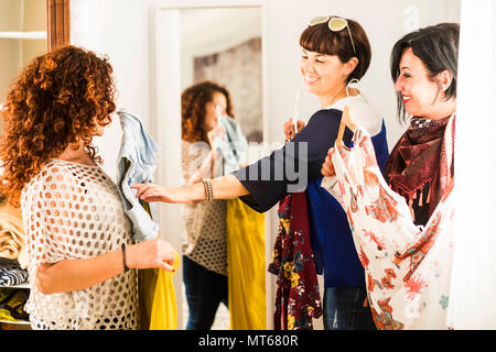 group of three caucasian friends choosing the right dress to go out for a event or shopping leisure outdoor activity. home interior with a lot of dres Stock Photo