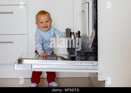 Toddler child, boy, helping mom, putting dirty dishes in dishwasher at home, modern kitchen Stock Photo