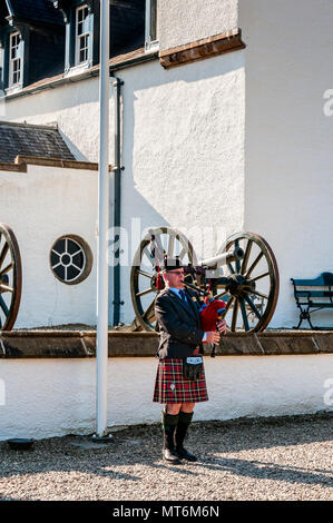 A Scottish piper in traditional highland dress plays typical airs on the bagpipes standing before a wall outside the tall white façade of Blair Castle Stock Photo