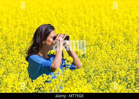 Young woman looking through binoculars in flowering yellow rapeseed field Stock Photo